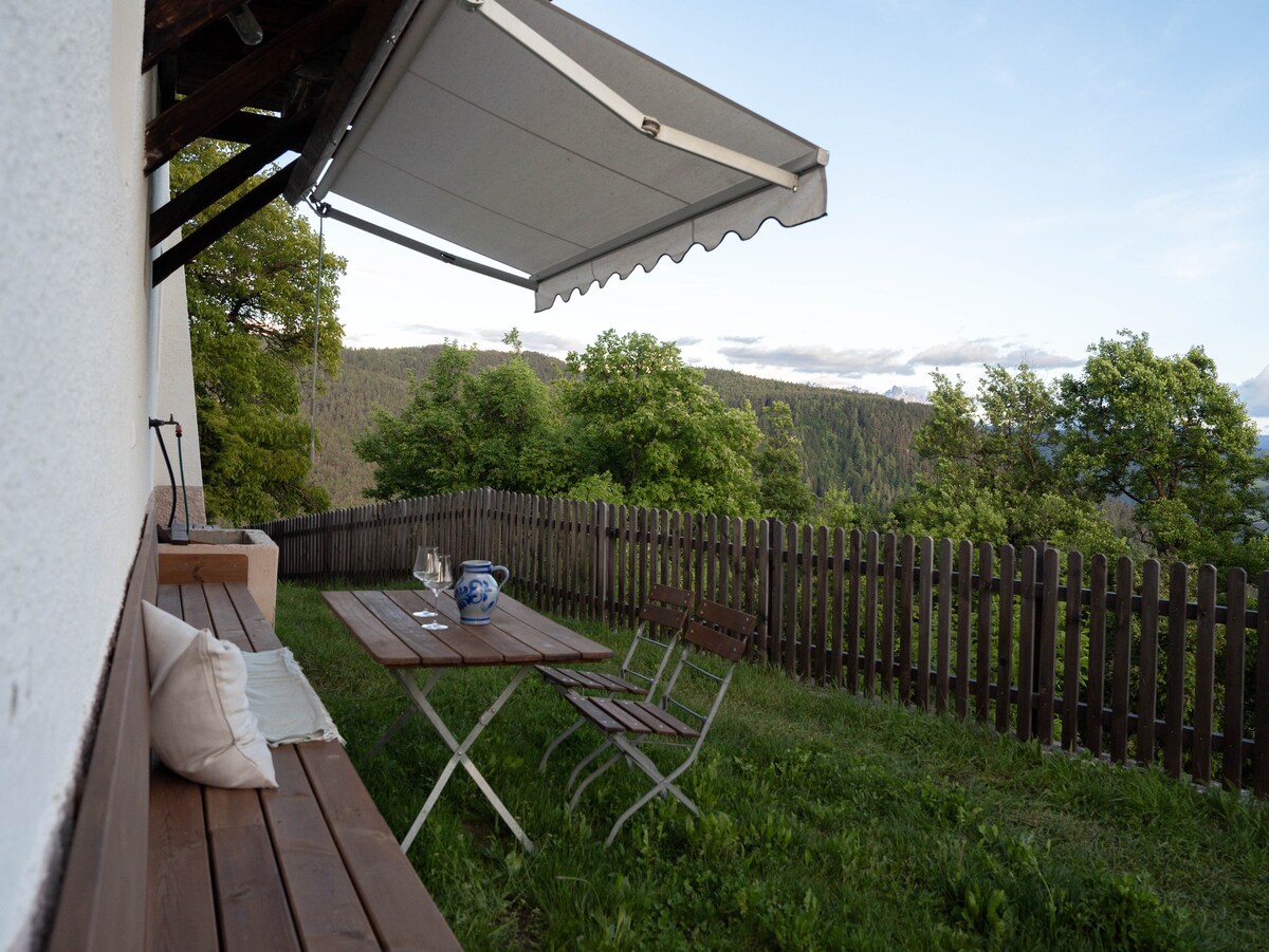 A shaded outdoor terrace features a wooden table and folding chairs, set against a backdrop of lush greenery and rolling hills. A protective awning extends over the seating area, providing comfort for outdoor dining or relaxation. The surrounding fence enhances the private setting.