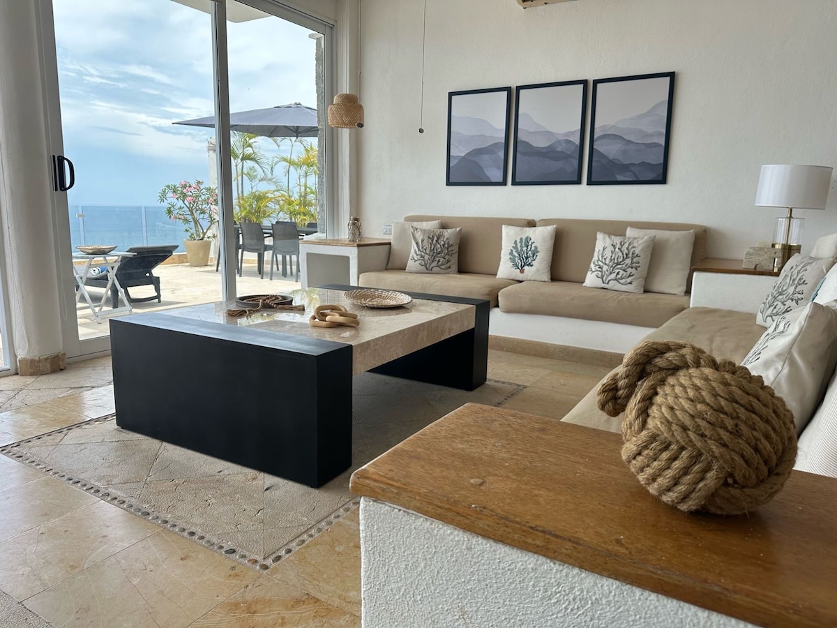 A bright and airy living room is portrayed, featuring a spacious sectional sofa adorned with decorative cushions. A coffee table with a simple design sits at the center. Large glass doors provide access to a terrace with outdoor seating and scenic views.