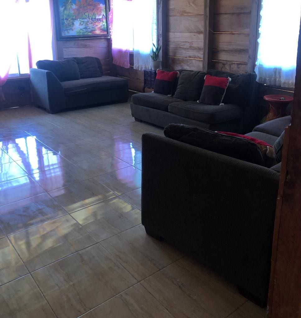 A spacious living area features two comfortable sofas adorned with decorative cushions. Natural light is allowed to enter through large windows draped with light curtains, illuminating the polished tile floor and wooden wall accents.