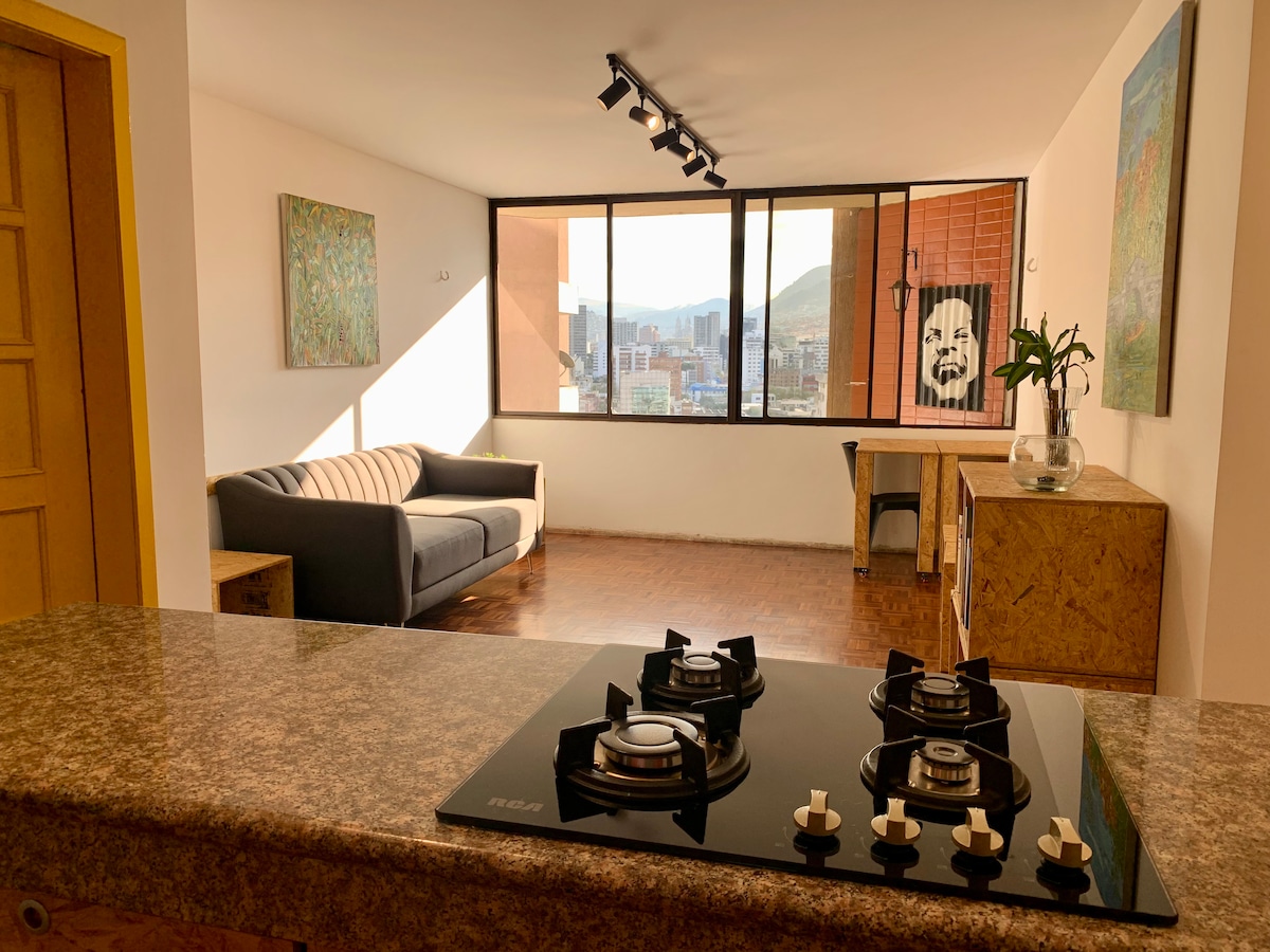 A bright living space features a comfortable sofa placed beside large windows offering views of the city. A modern kitchen area is visible in the foreground, with a sleek stove on a granite countertop. Natural light enhances the inviting atmosphere.