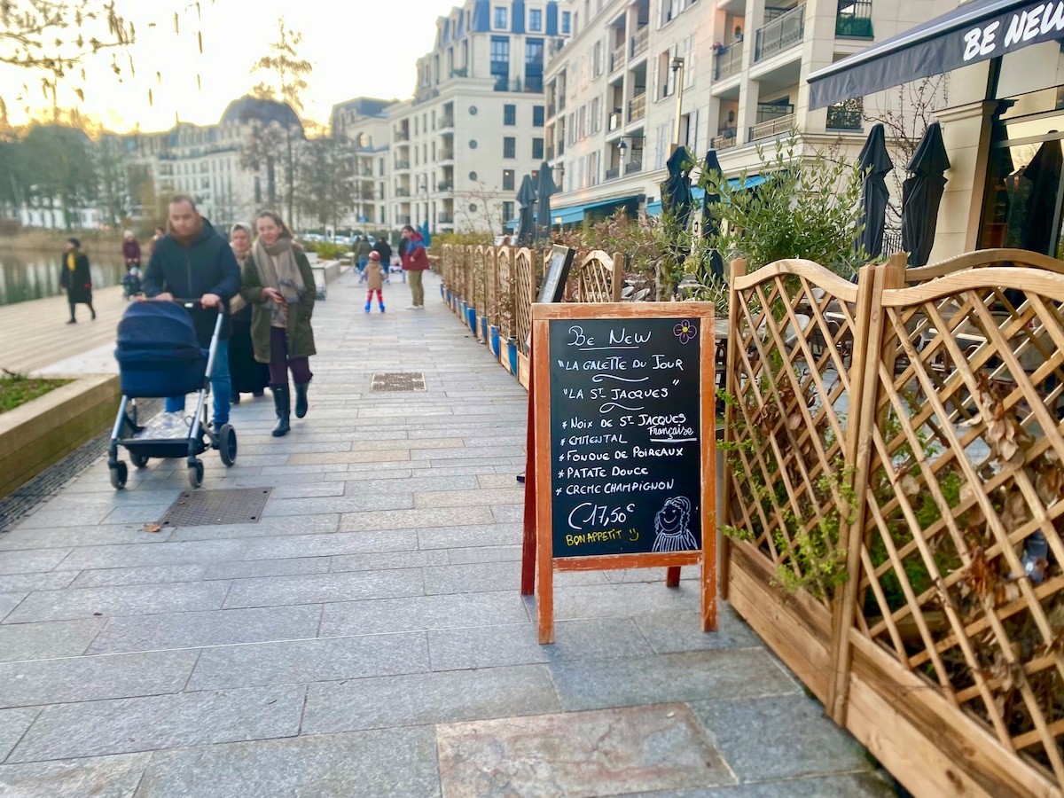 A chalkboard sign stands along a pedestrian pathway, displaying daily specials. A wooden trellis lines the path, providing a decorative element. People stroll by, with one pushing a stroller, and café seating is visible in the background, creating a lively atmosphere.