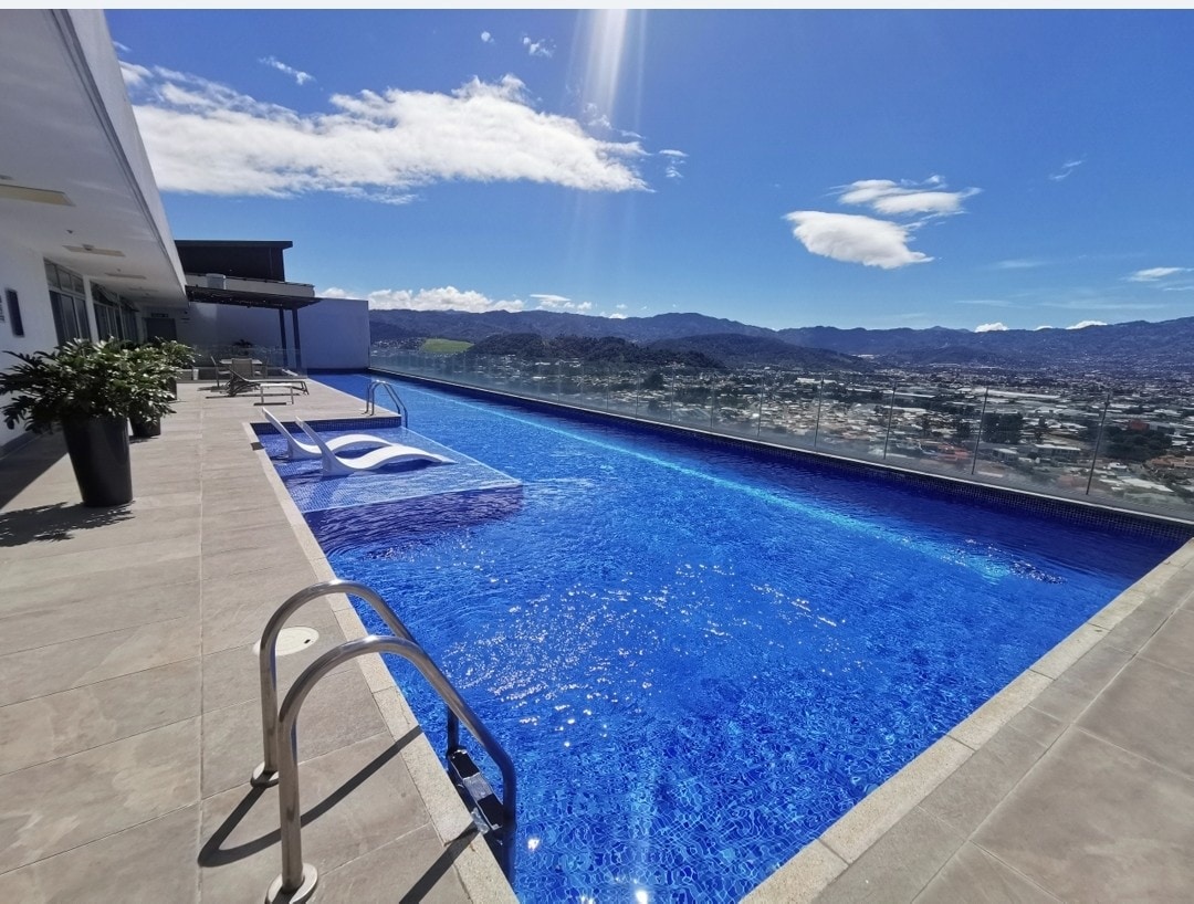 An expansive infinity pool stretches along the terrace, offering views of the surrounding city and mountains. Sun loungers are positioned nearby, and a bright blue sky is reflected in the water, creating a serene atmosphere.