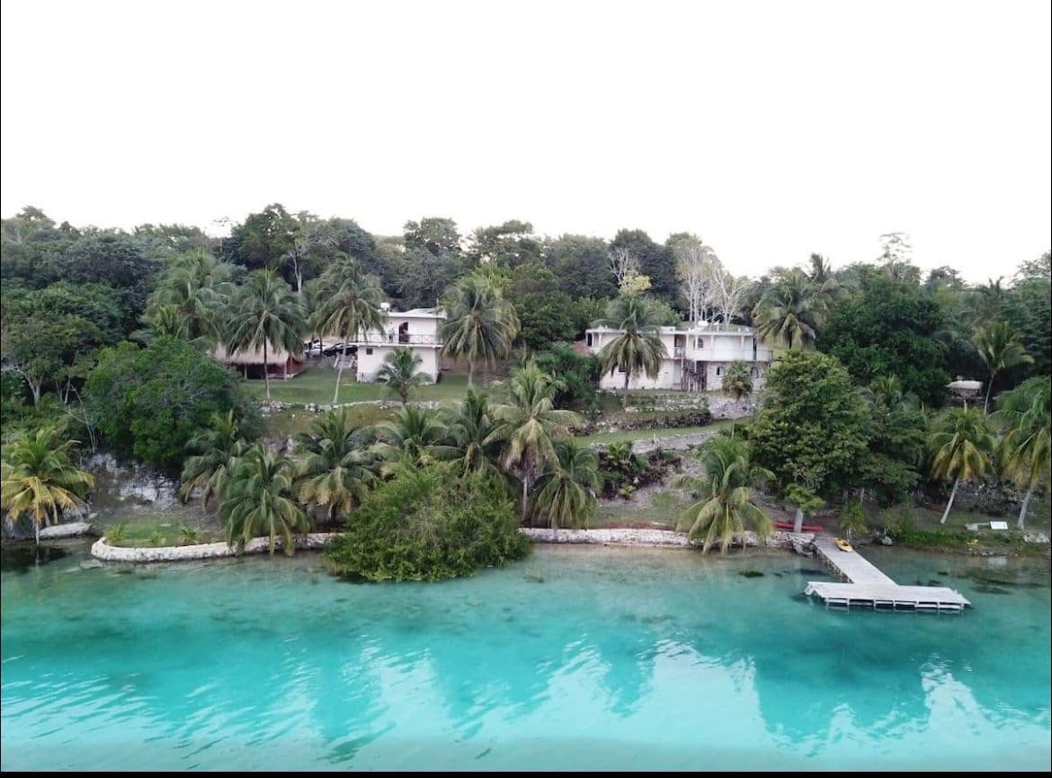 A serene waterfront property is surrounded by lush greenery and palm trees. The clear turquoise water gently meets the shoreline, with a wooden dock extending into the water. The landscape is complemented by a spacious lawn area and multiple buildings nestled among the trees.