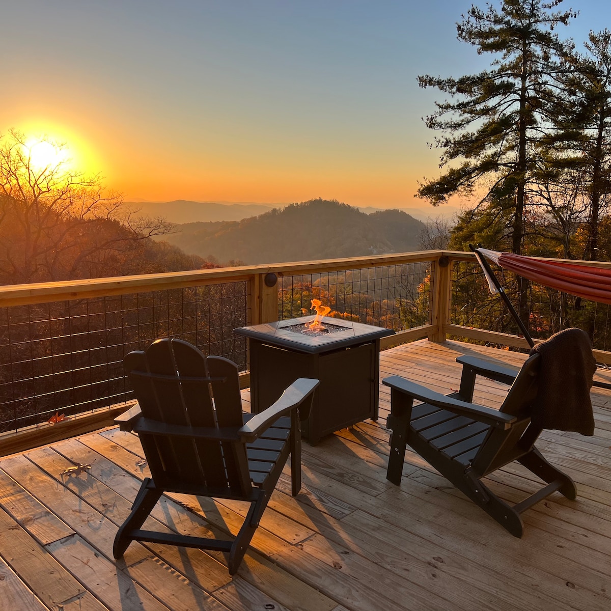 A wooden deck is viewed at sunset, featuring two black Adirondack chairs positioned around a fire table. A hammock is visible, and the landscape boasts distant mountains with a warm glow from the setting sun.