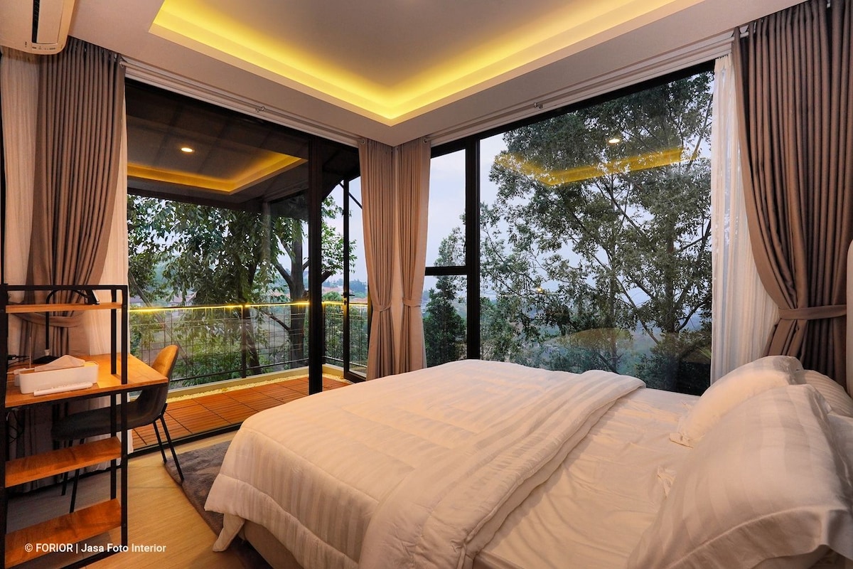 A cozy bedroom features a spacious bed with soft linens and a view through large glass windows. The room is complemented by neutral tones, and a small dining area with sleek chairs is visible. Natural light filters in, enhancing the connection to the outdoor scenery.