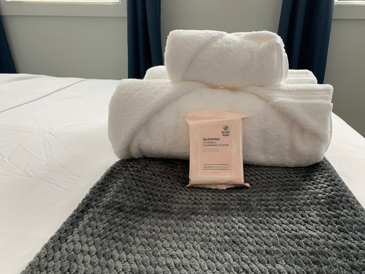 Soft, white towels are neatly stacked on a made bed, accompanied by a pack of cleansing cloths placed on a textured gray mat. Natural light filters through nearby windows, highlighting the simplicity and freshness of the linens.