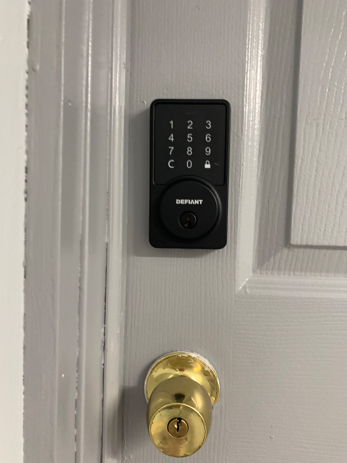 A modern door lock is displayed on a white door. The keypad features numbered buttons from 1 to 9, with a prominent circular keyhole positioned below. A shiny gold doorknob is visible beside the lock, contrasting with the door's matte finish.