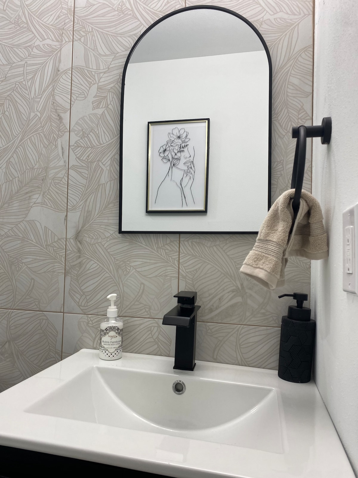 A modern bathroom features a compact white sink mounted against patterned wall tiles. A curved mirror is framed in black, reflecting an artistic print above. A hand soap dispenser and a dark-colored lotion pump are positioned beside the sink, with a towel hanging from a wall-mounted holder.