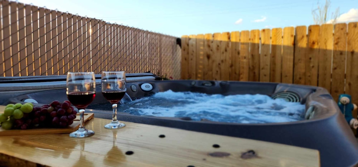 A private hot tub fills the image, featuring bubbling water and accessible controls. Two glasses of red wine rest on an adjacent wooden platform, accompanied by a wooden platter of mixed grapes. A wooden fence provides privacy while blue sky and light clouds are visible overhead.