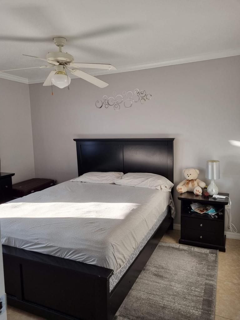 A spacious bedroom features a dark wooden bed frame with a light-colored bedding set. A bedside table holds a lamp and a teddy bear. Soft light is provided by a ceiling fan, and a decorative wall piece complements the neutral color scheme.