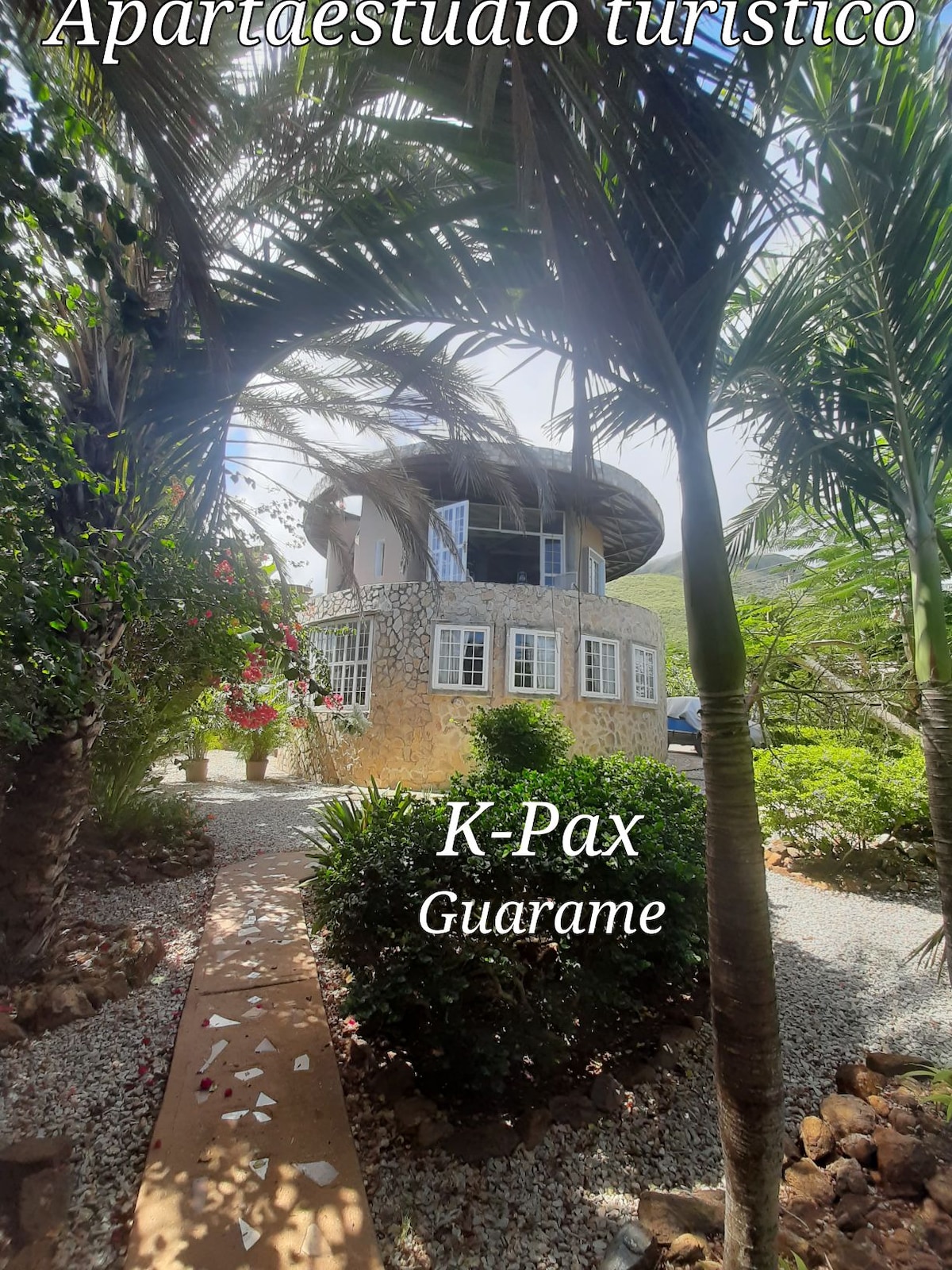 A round apartment building is surrounded by lush greenery and tropical plants. A stone pathway leads towards the entrance, framed by vibrant flowers. The structure showcases a mix of stone and glass, offering a harmonious integration with the natural landscape.