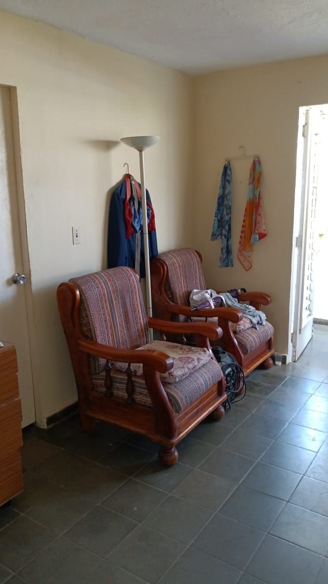 Two wooden armchairs with patterned upholstery are positioned beside a door that opens to the outside. A standing lamp stands nearby, and clothes hang from wall hooks, adding a touch of functionality to the space. The floor is covered in dark, square tiles.