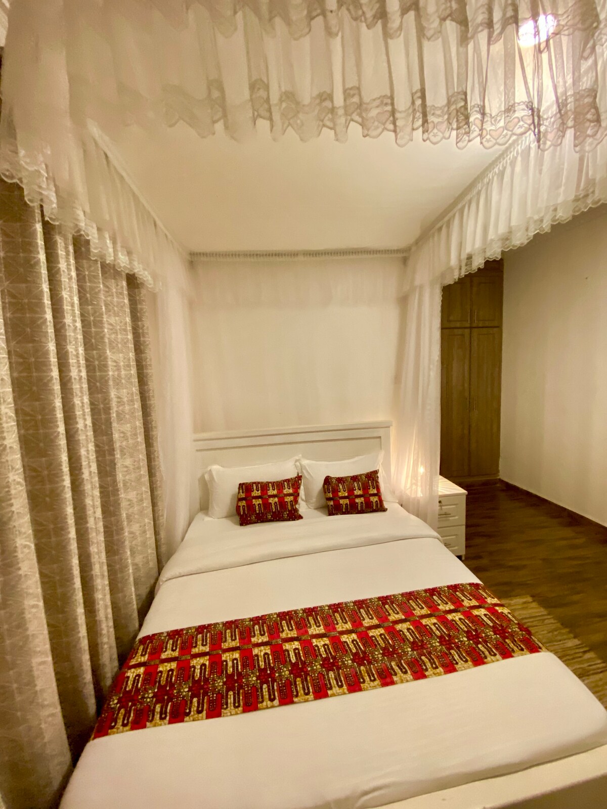 A spacious bed is dressed in crisp white linens with decorative cushions featuring vibrant patterns. Sheer curtains softly frame the bed, allowing natural light to fill the room. A wooden floor and a sleek nightstand contribute to the room's simple elegance.