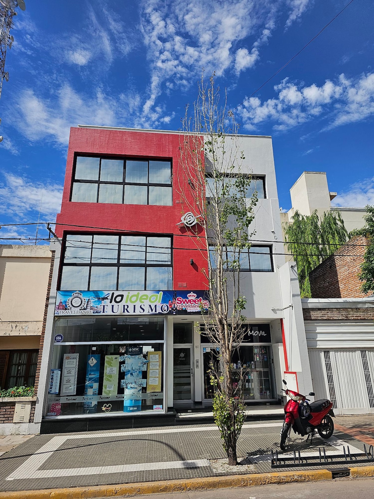 The exterior view of a three-story building displays large glass windows on the upper floors. The facade features a combination of red and gray paint. A small tree is positioned in front, while a motorcycle is parked nearby along the sidewalk.