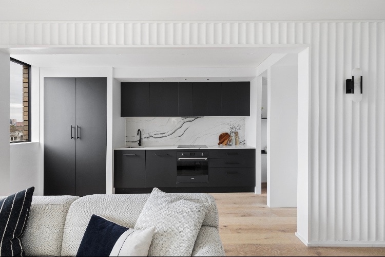 A modern kitchen space features sleek black cabinetry complemented by a marble backsplash. Natural light fills the area through large windows, highlighting the open layout that connects to a comfortable living space with textured cushions on a light-colored sofa.