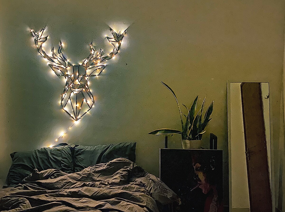 A cozy bedroom is highlighted by a decorative light installation shaped like a stag's head on a muted wall. A spacious bed with rumpled bedding is positioned in front, while a potted plant adds greenery to the space. A tall mirror leans against the wall, reflecting the surroundings.