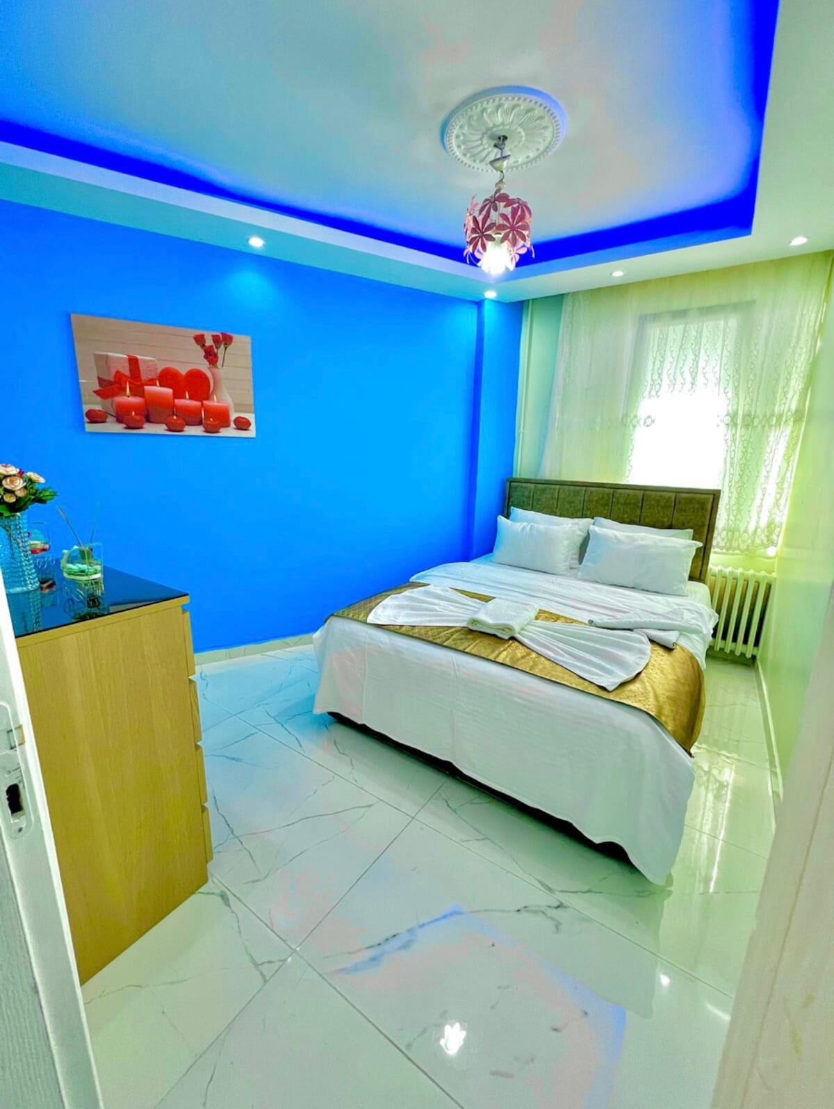 A bright and inviting bedroom is presented, featuring a spacious bed with decorative linens. A vibrant blue wall adds a pop of color, while a chandelier hangs from the ceiling. Natural light filters through sheer curtains, illuminating the room's clean surfaces.