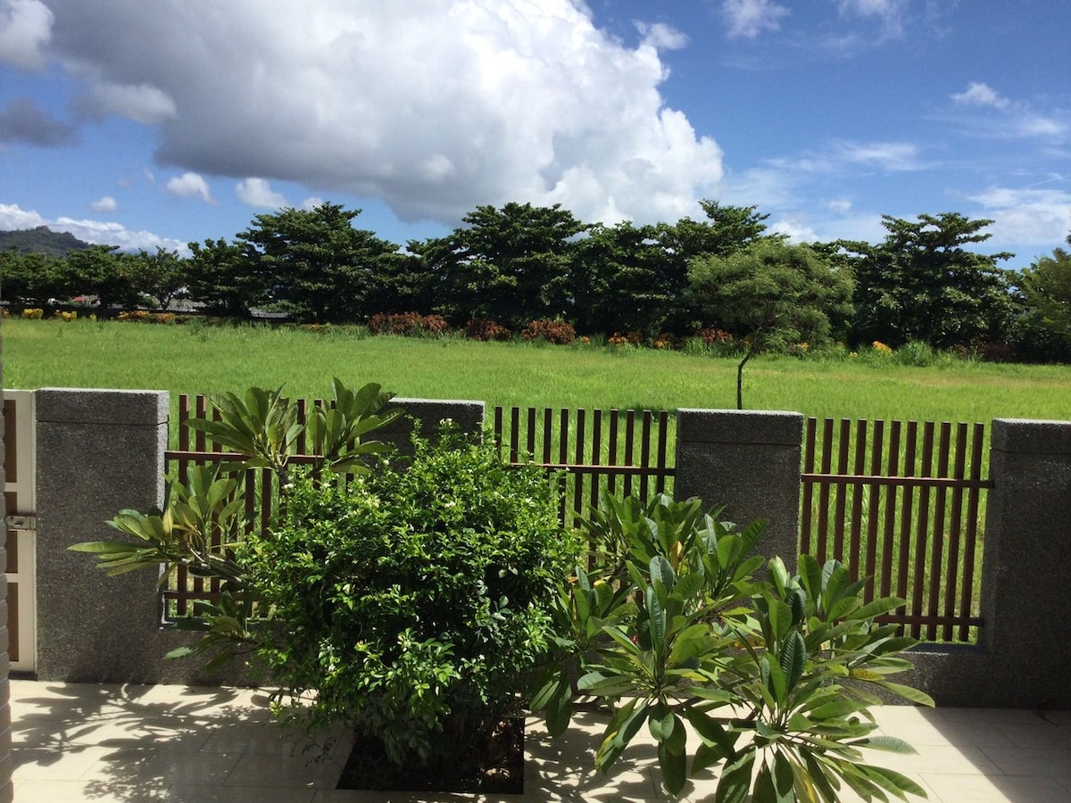 A view of a lush green field is framed by a wooden fence and complemented by vibrant foliage in the foreground. The sky is partly cloudy, with bright sunlight illuminating the scene.