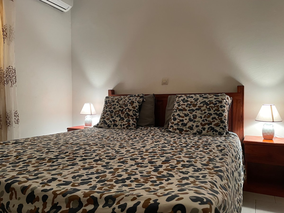 A cozy bedroom features a spacious bed layered with a patterned duvet and matching pillows. Soft bedside lamps illuminate the room, creating a soothing ambiance. Light-colored walls and tiled flooring enhance the sense of openness and tranquility.
