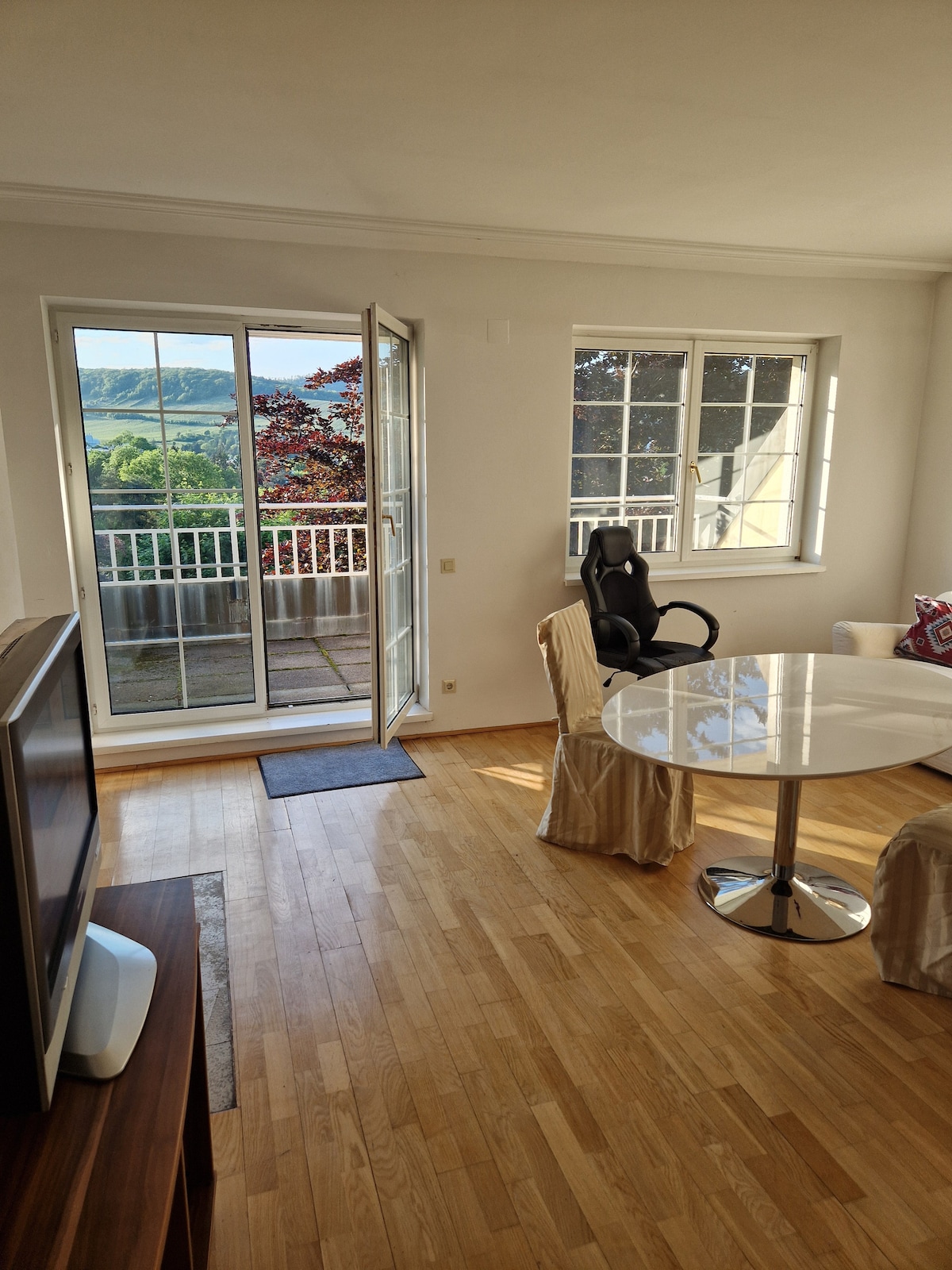 A bright living area features large windows, inviting natural light and a view of the outdoors. A round table sits at the center, accompanied by a modern chair. An open door leads to a patio area, enhancing the connection to the outside scenery.