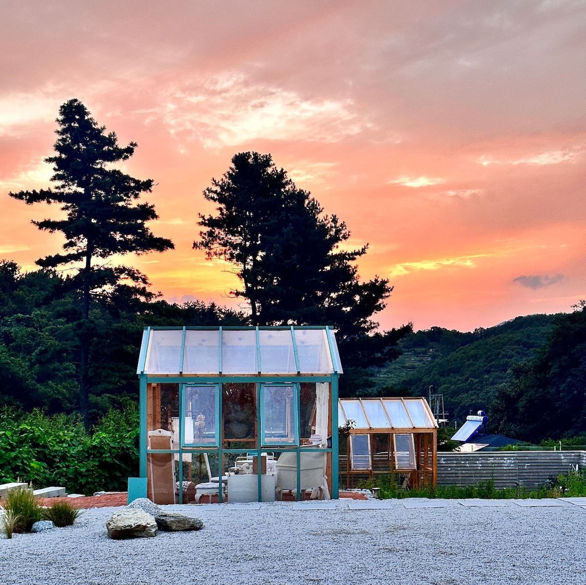 Two greenhouses are positioned against a colorful sky at sunset, framed by tall trees and lush greenery. Their glass panels reflect the warm light, while stone paths lead toward them, inviting exploration in this peaceful outdoor setting.