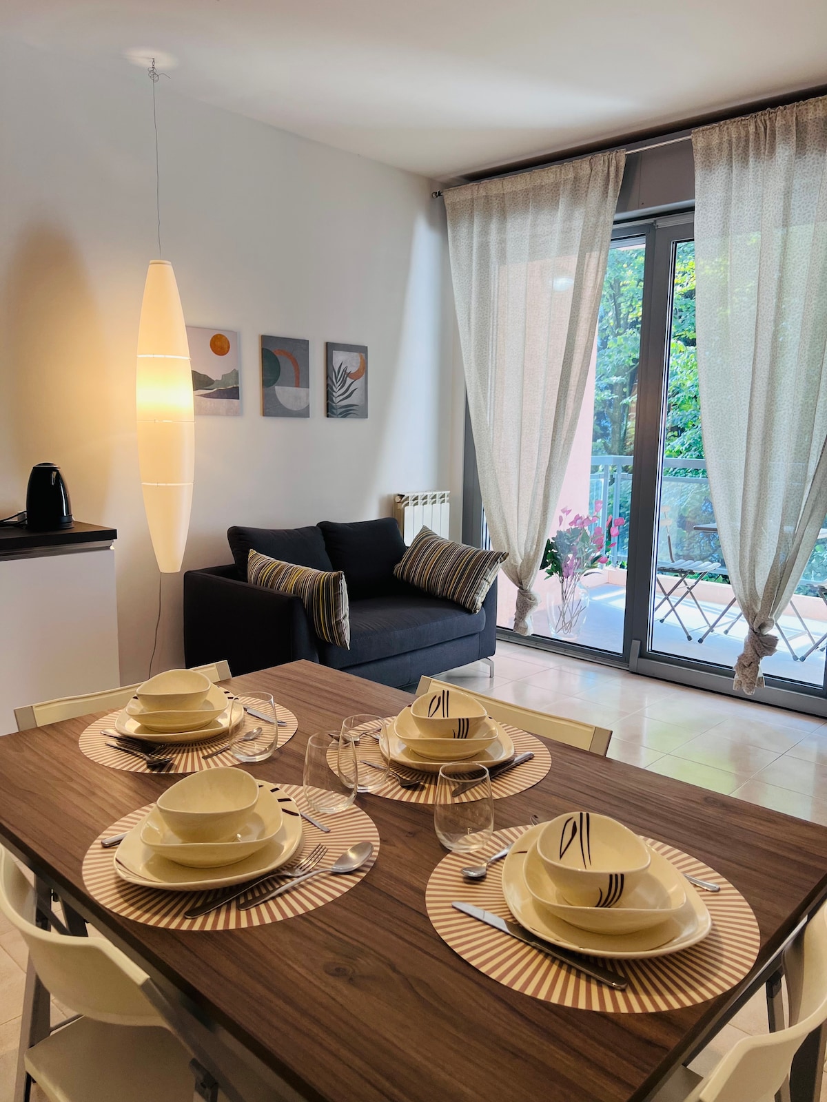 The dining area features a wooden table set for four, complete with dishware and cutlery. A dark sofa with patterned cushions rests nearby, while large glass doors offer natural light and a view of the terrace, framed by flowing sheer curtains.