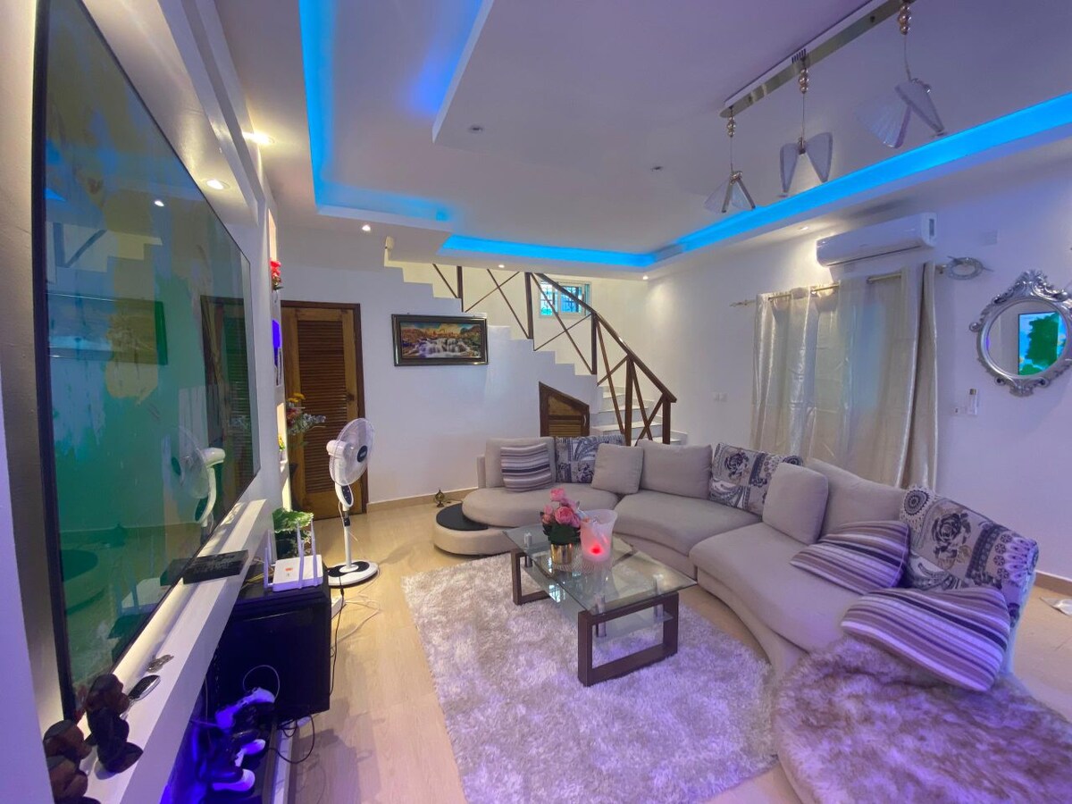 A modern living room features a curved sectional sofa adorned with decorative cushions. Soft lighting highlights a low coffee table and an entertainment unit with a large screen. A staircase is visible in the background, complemented by light-colored walls and a plush area rug.