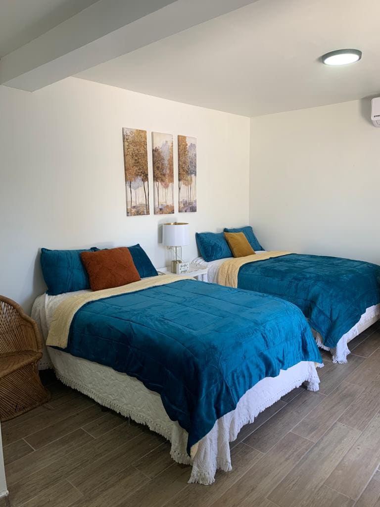 A serene sleeping area features two spacious beds topped with teal bedspreads and accented by colorful pillows. A soft throw blanket drapes over one bed. A bedside table with a lamp is positioned between the beds, and three nature-themed artworks adorn the wall.