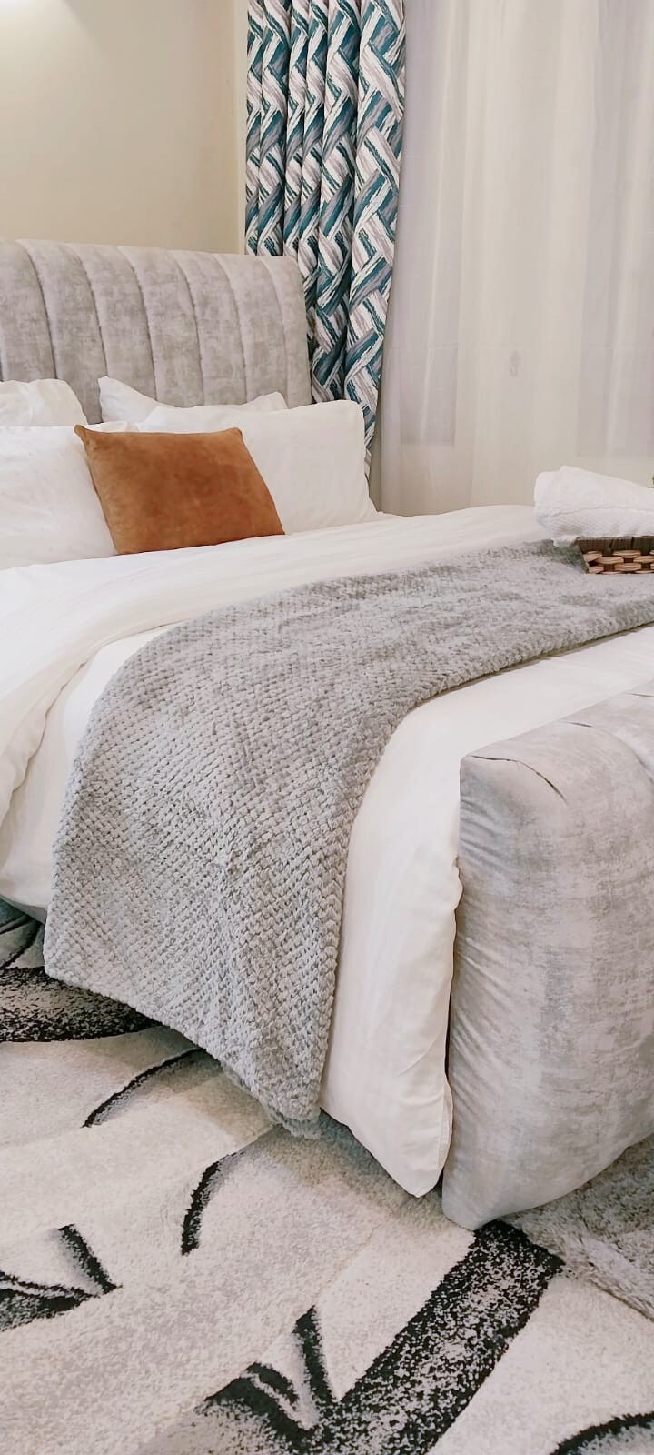 A spacious bed is dressed in soft white linens and layered with a textured grey throw. A rust-colored pillow provides a warm accent. The headboard features a plush, tufted design. A patterned rug complements the overall aesthetic of the room.