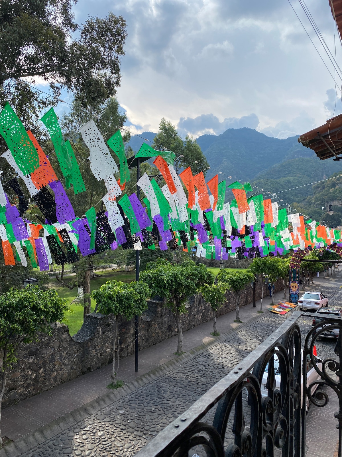 A vibrant display of papel picado in various colors hangs across the street, adding character to the scene. Lush trees and a stone wall frame the cobblestone road below, while rolling mountains rise in the background under a partly cloudy sky.