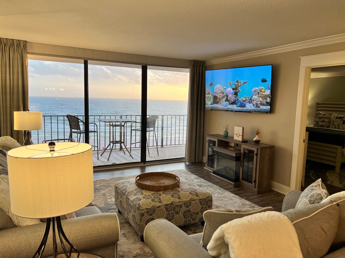 The living room features large glass doors that offer breathtaking ocean views and access to the private balcony. A flat-screen TV is mounted on the wall, while comfortable seating arrangements are provided. Natural light floods the space through the windows, creating a bright atmosphere.
