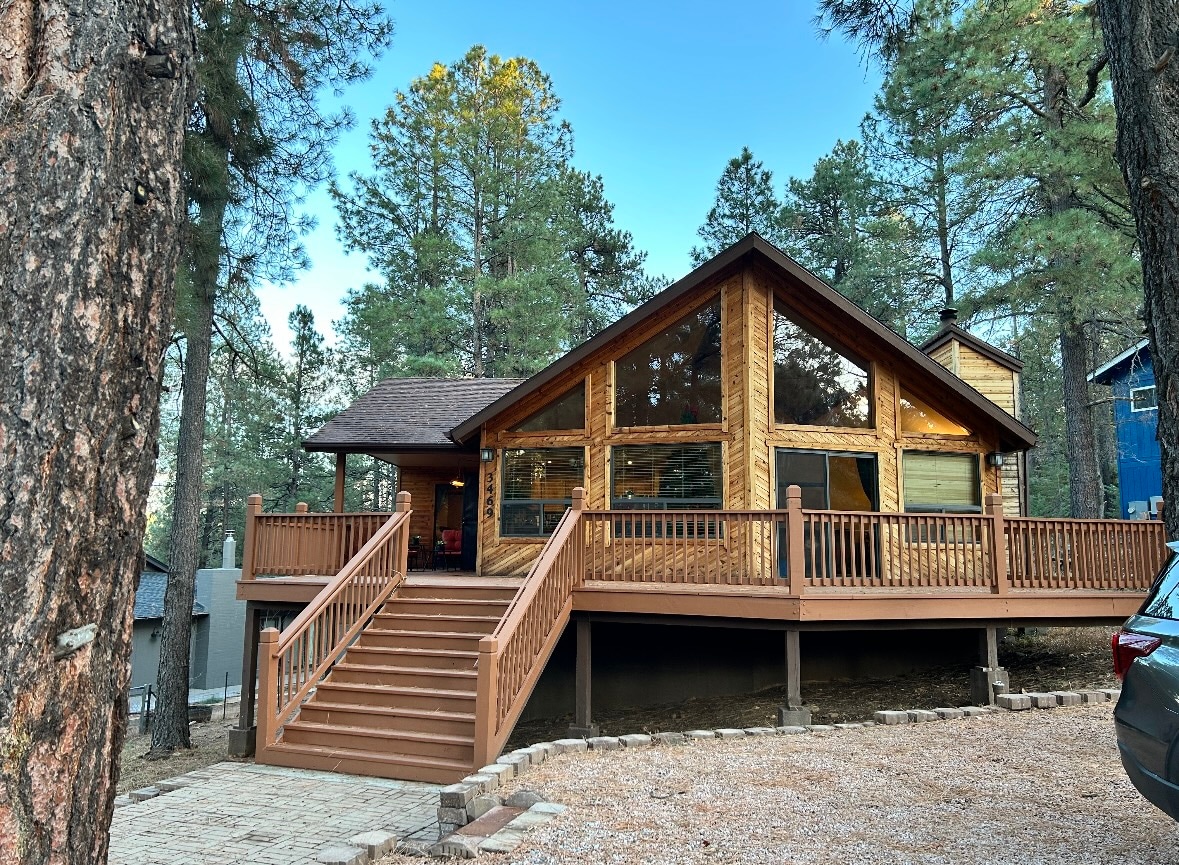 A cozy cabin exterior showcases large windows that invite natural light, surrounded by tall pine trees. A spacious deck with railing leads to the front entrance, complemented by a well-maintained gravel area and a driveway for parking.