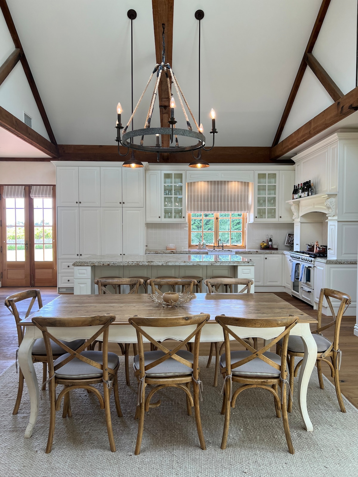 A spacious kitchen is featured with high ceilings and wooden beams. A large wooden dining table is surrounded by multiple crossback chairs. White cabinetry and a marble countertop enhance the aesthetic, while large windows allow natural light to fill the room.