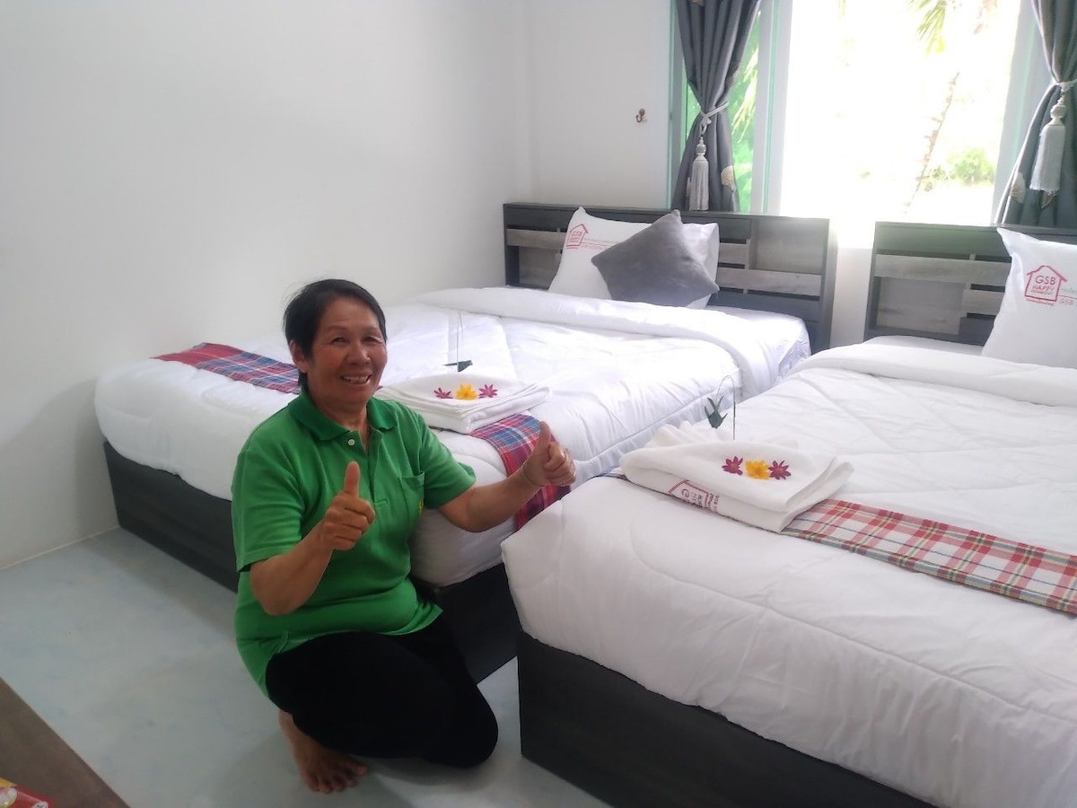 A well-lit room features two spacious beds dressed in crisp white linens. Decorative towels and fresh flowers are arranged on each bed. Soft curtains frame a window that allows natural light to brighten the space, creating a welcoming atmosphere.