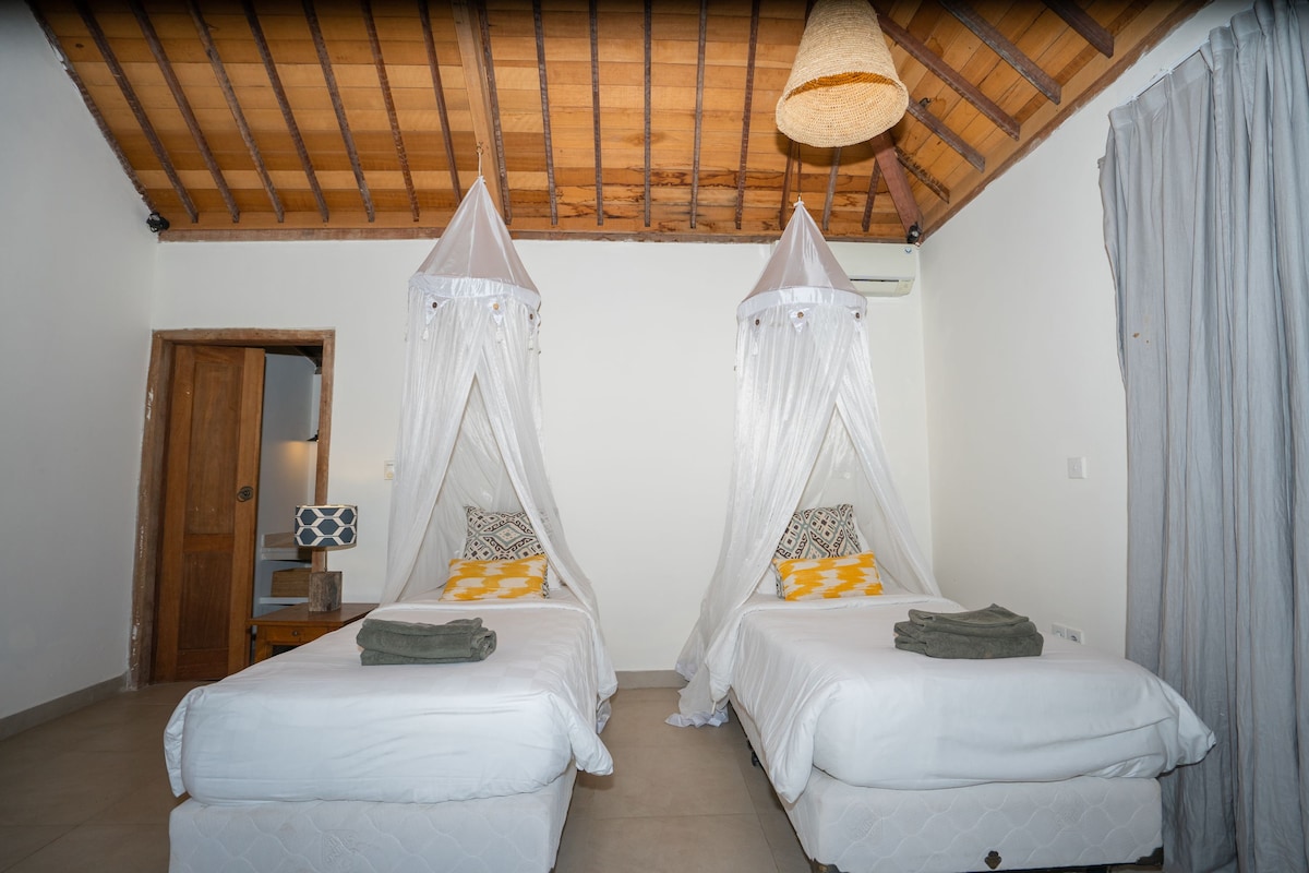 The room features two single beds, each draped with light fabric canopies. Fresh towels are neatly arranged on the beds, which are set against a light-colored wall. A simple nightstand and a woven lamp add warmth to the space, with soft natural light illuminating the room.