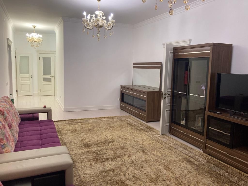 A spacious living room features elegant lighting fixtures hanging from a high ceiling. A plush sofa is situated on a warm-toned rug, complemented by a modern entertainment unit and glass-fronted cabinets. The room is brightened by neutral walls and natural light from nearby doors.