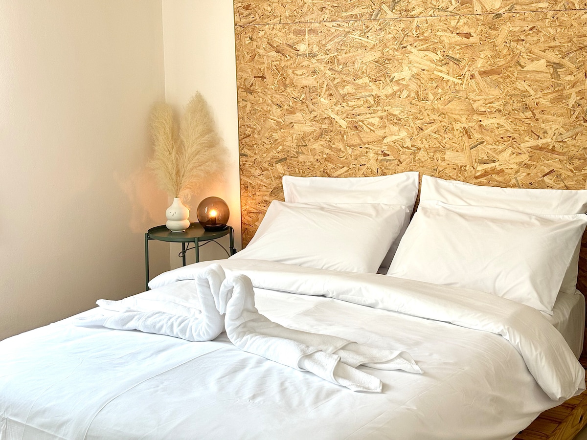 A spacious bed with neatly arranged white linens is complemented by a decorative wooden wall. A small table holds a soft glow lamp and a vase with pampas grass, adding a hint of warmth to the serene space.