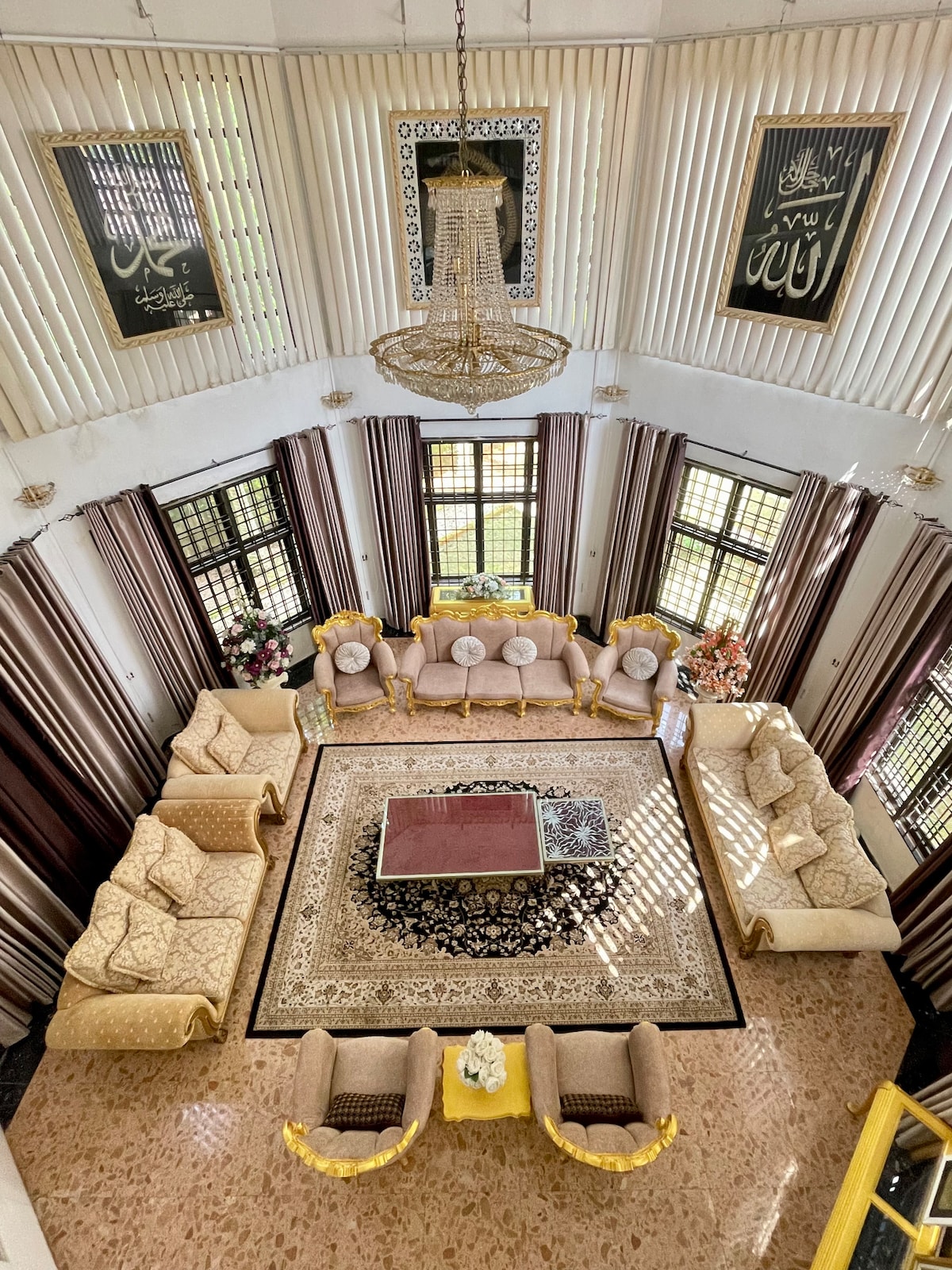 A spacious living area is arranged with multiple seating arrangements featuring light-colored upholstery. A decorative rug lies at the center, accompanied by a polished table. Floor-to-ceiling windows fill the room with natural light, complemented by patterned curtains.