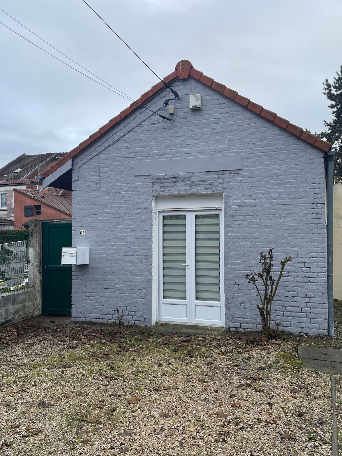 The exterior of a small, grey house showcases a pitched roof with a red-tiled edge. A white double door leads to the entrance, surrounded by a gravelled area. Nearby, a green gate and wall can be seen, alongside sparse vegetation.