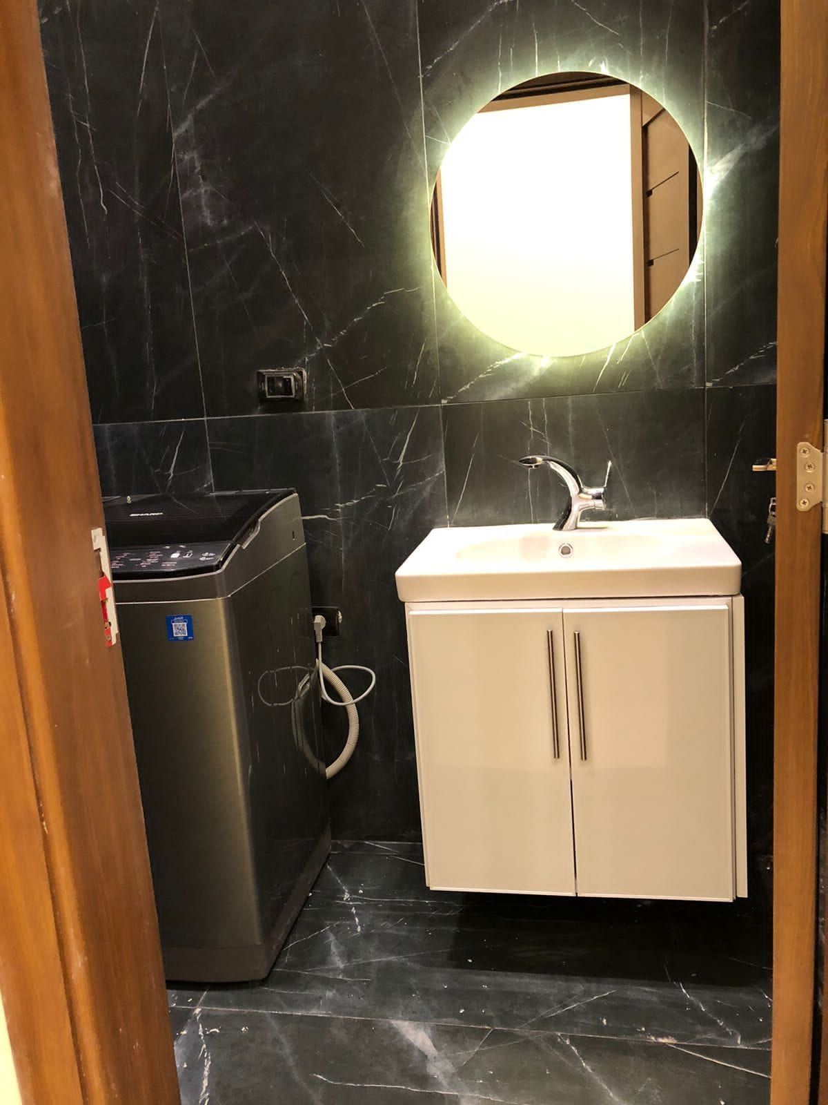 A well-lit bathroom features a round mirror above a modern sink with two cabinet doors offering storage space. A compact washing machine is positioned beside the sink, against a backdrop of dark, marbled wall tiles that enhance the contemporary design.