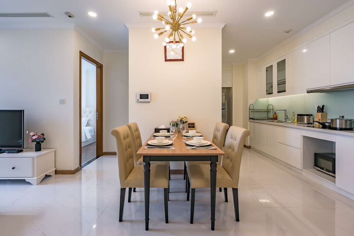 A dining area features a rectangular table surrounded by six upholstered chairs. A modern light fixture hangs from the ceiling, providing soft illumination. The kitchen is visible in the background, equipped with white cabinetry and modern appliances, while a television rests on a console nearby.