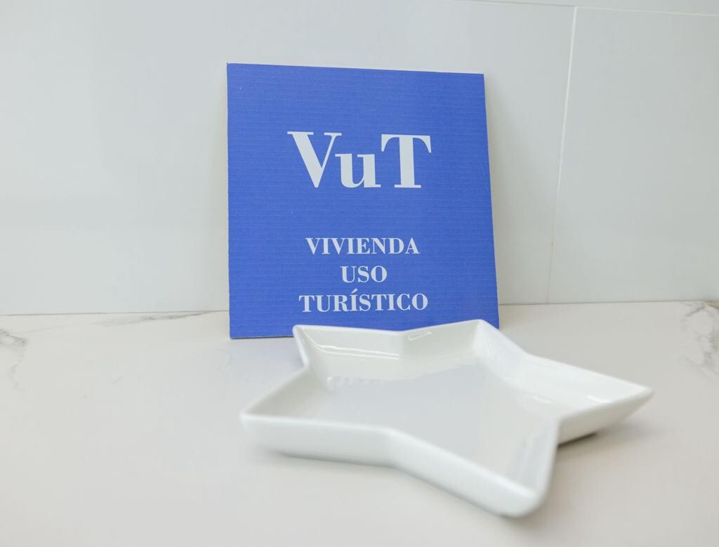 A blue sign displaying 'VuT VIVIENDA USO TURÍSTICO' is positioned next to a white star-shaped dish. The minimalistic design emphasizes the welcoming nature of the accommodation, set against a light background.