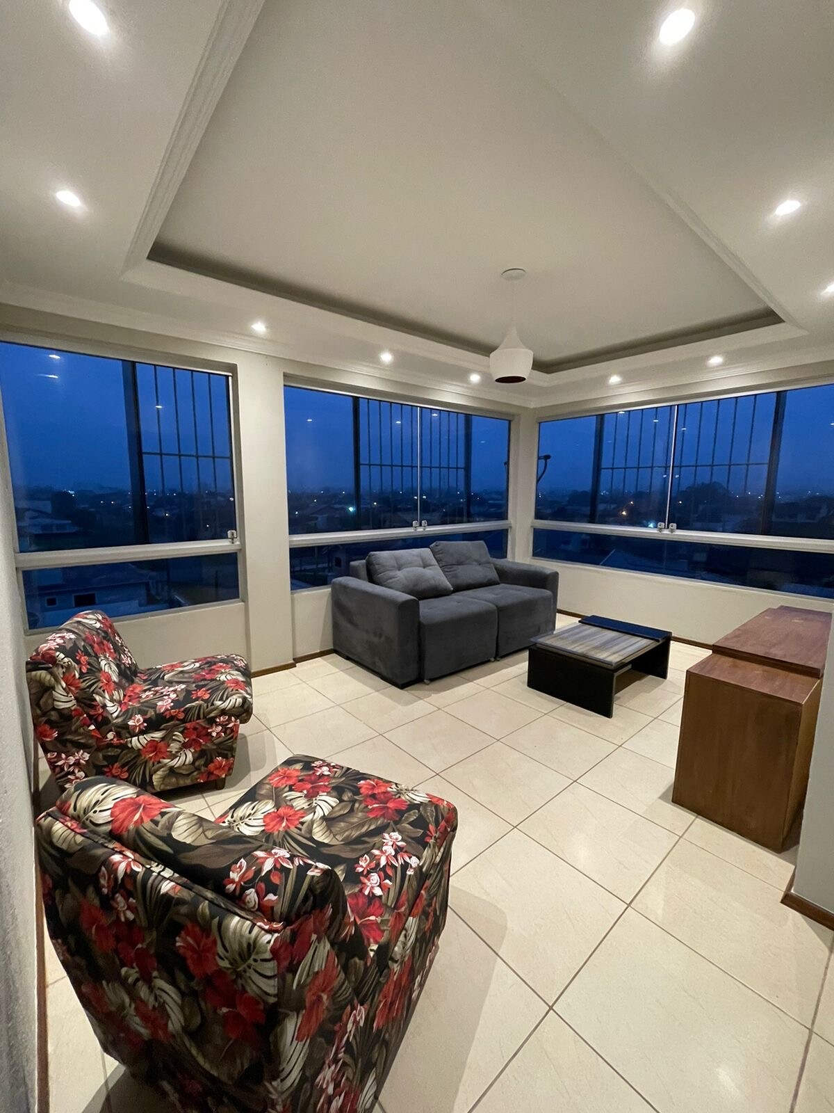 A bright living area features large windows that offer expansive views of the surroundings. Comfortable floral chairs are arranged alongside a gray sofa and a coffee table. Soft lighting highlights the geometric ceiling design, creating an inviting space for relaxation.