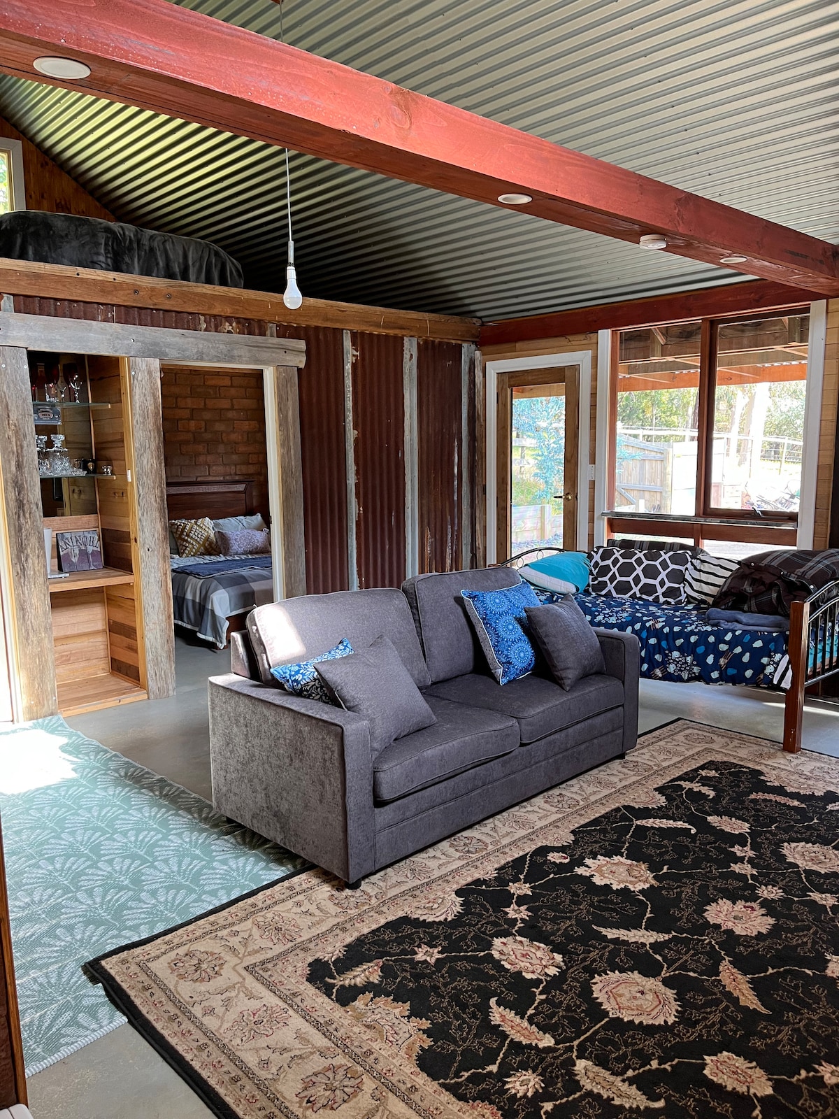 A cozy seating area features a dark gray couch with blue accent pillows. A decorative area rug overlays the concrete floor, and large windows allow natural light to illuminate the space. A wooden shelf with drinks can be seen in the background, enhancing the rustic charm.