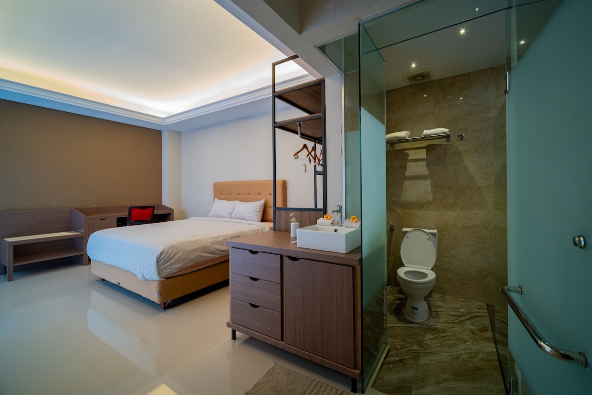 A well-lit room features a spacious bed against a neutral wall. A modern dresser and a small seating area are present. To the side, a glass-enclosed bathroom is visible, showcasing contemporary fixtures and soft lighting.