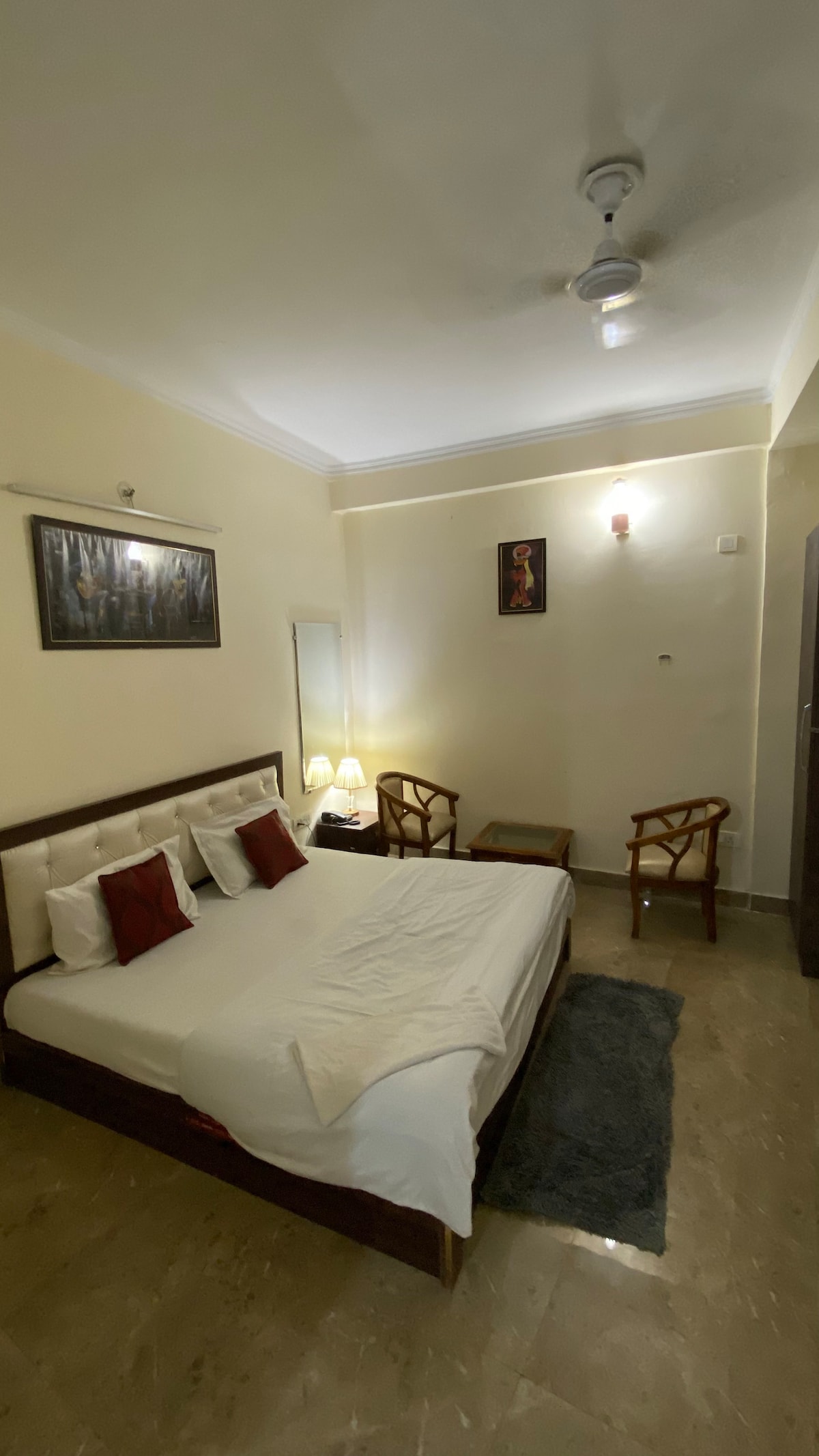 A comfortable sleeping area features a spacious bed with white linens and decorative pillows. Two armchairs are positioned beside the bed, with a mirror reflecting soft lighting. Wall art hangs above the bed, adding visual interest to the neutral-colored room.