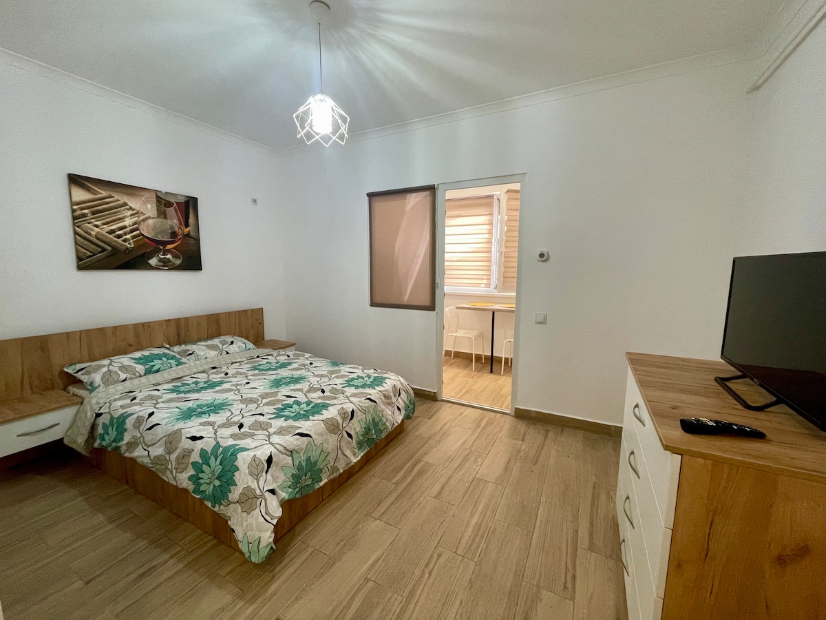 A spacious bedroom features a double bed with a floral-patterned blanket and a light fixture overhead. A wooden dresser is positioned next to the bed, while a television is displayed on a console. A doorway leads to a well-lit area beyond.