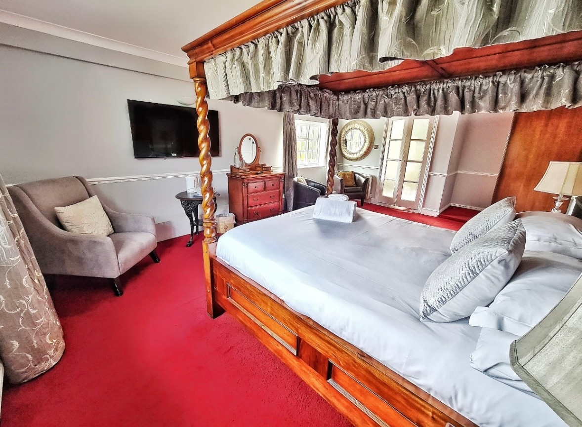 A spacious room features a superking four-poster bed with elegant bedding. A cozy armchair is positioned nearby, accompanied by a wooden dresser. A flat-screen TV is mounted on the wall, and double glass doors lead to additional light from outside.