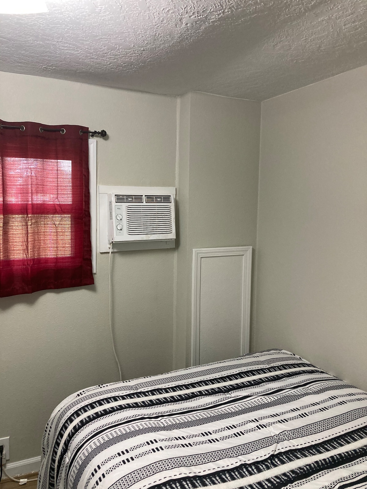 A cozy private room features a spacious bed covered with a striped blanket. A window draped in red curtains allows gentle light to enter, while a wall-mounted air conditioning unit provides cooling comfort.
