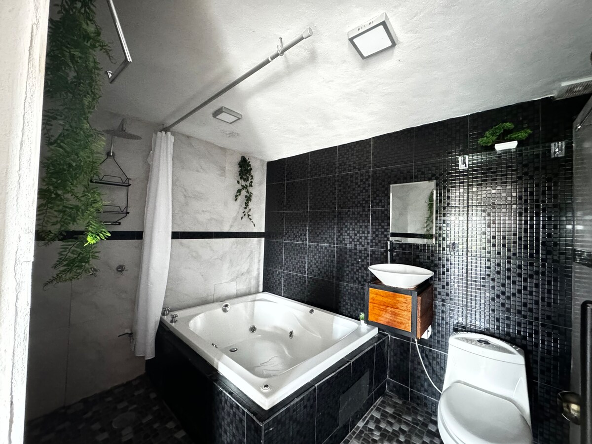 A modern bathroom features a spacious jacuzzi with hydromassage jets surrounded by stylish dark tiles. A simple wooden cabinet and a large mirror add to the design. Soft greenery hangs from the ceiling, providing a natural touch to the contemporary space.